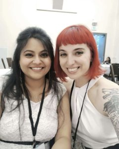 A close up of Meghalee and Rachael in a conference room smiling at the camera. Lanyards hang around their neck and they are both dressed in black and white.